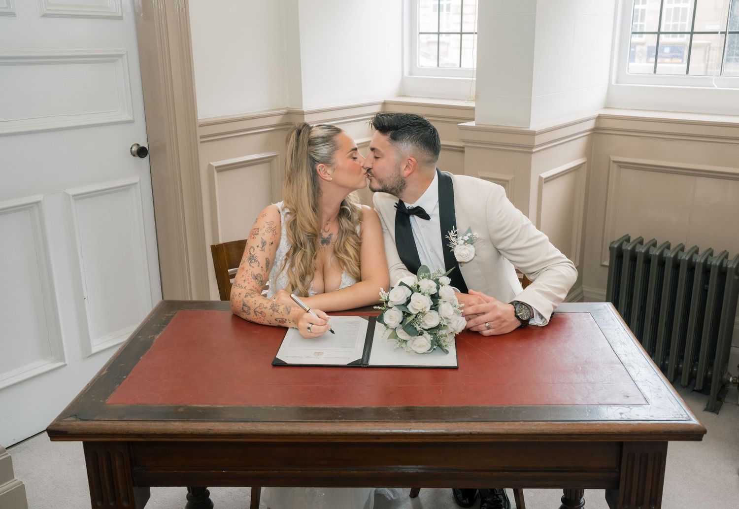 Newlywed couple kissing while signing wedding register at wooden desk in elegant ceremony room.