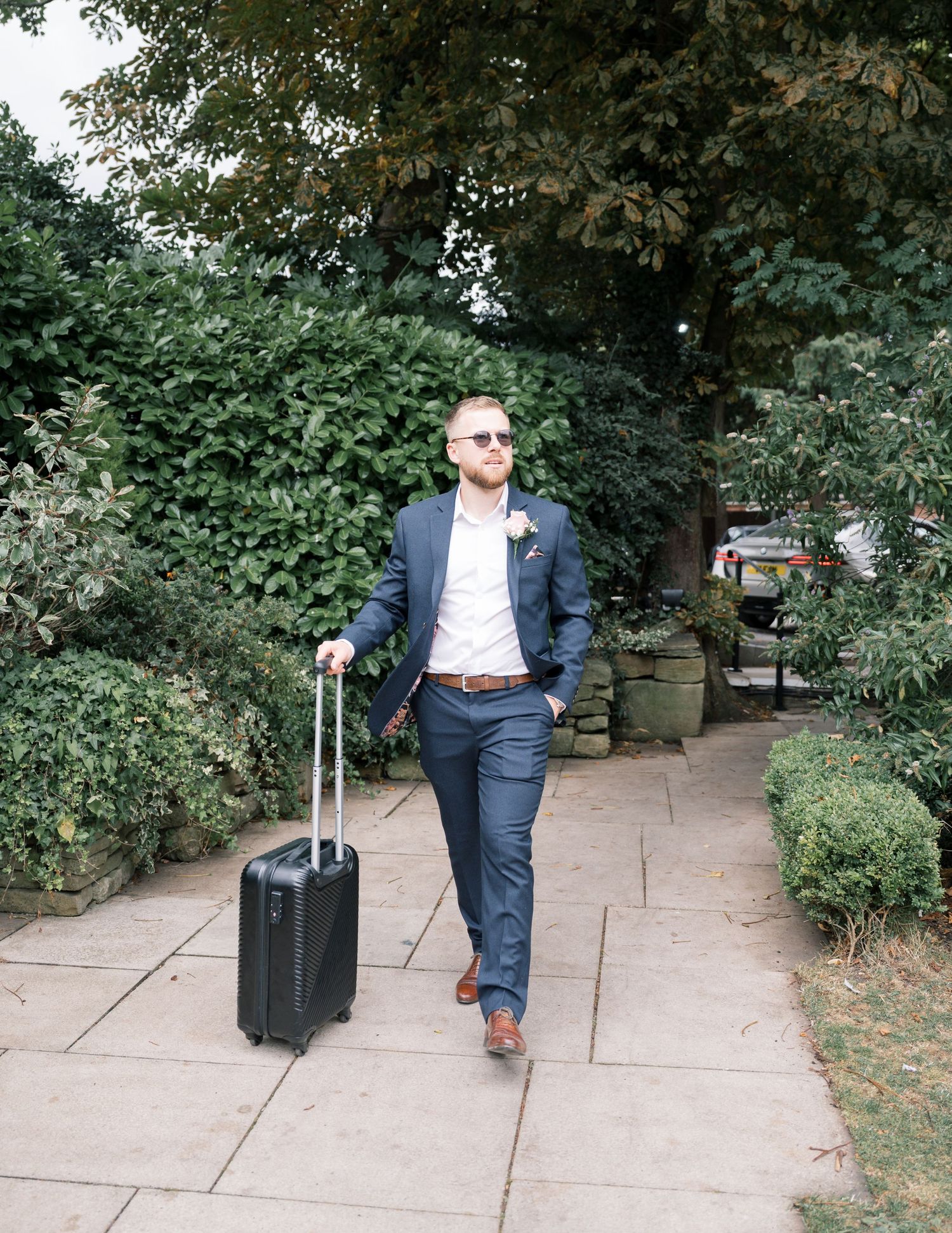 Person in navy suit walking along sidewalk pulling luggage through tree-lined path.