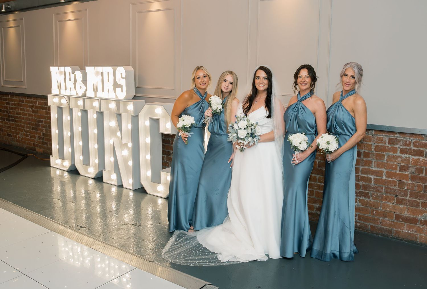 Bridesmaids in teal blue dresses posing with bride in front of illuminated marquee letters.