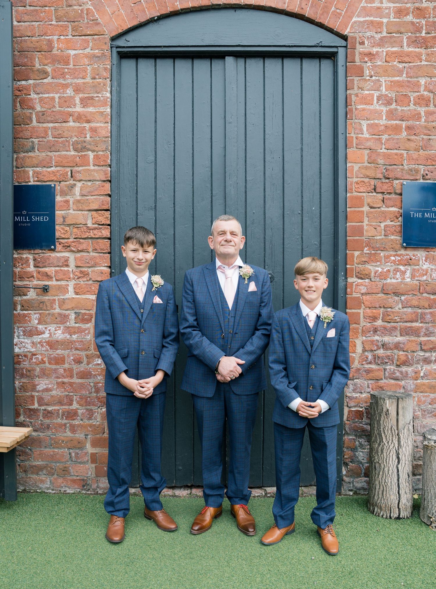 Groomsmen in navy suits and brown shoes posing against a brick wall with green turf.