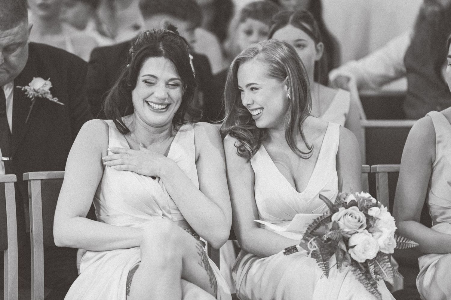 Two bridesmaids laughing joyfully during a wedding ceremony, wearing white dresses, captured in black and white.