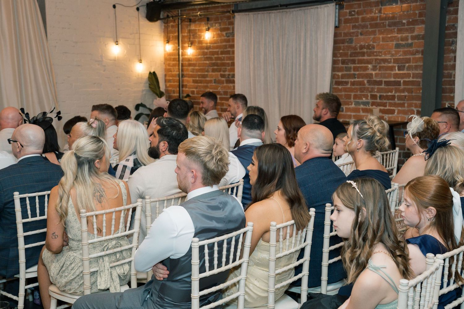 Wedding guests seated in white chairs watching ceremony in industrial brick venue with hanging lights.