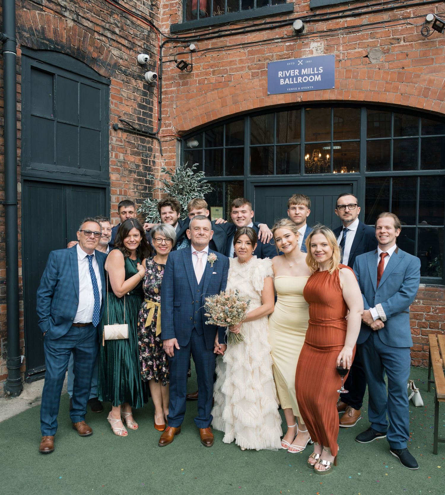 Wedding party poses for group photo outside brick industrial venue with blue doors.