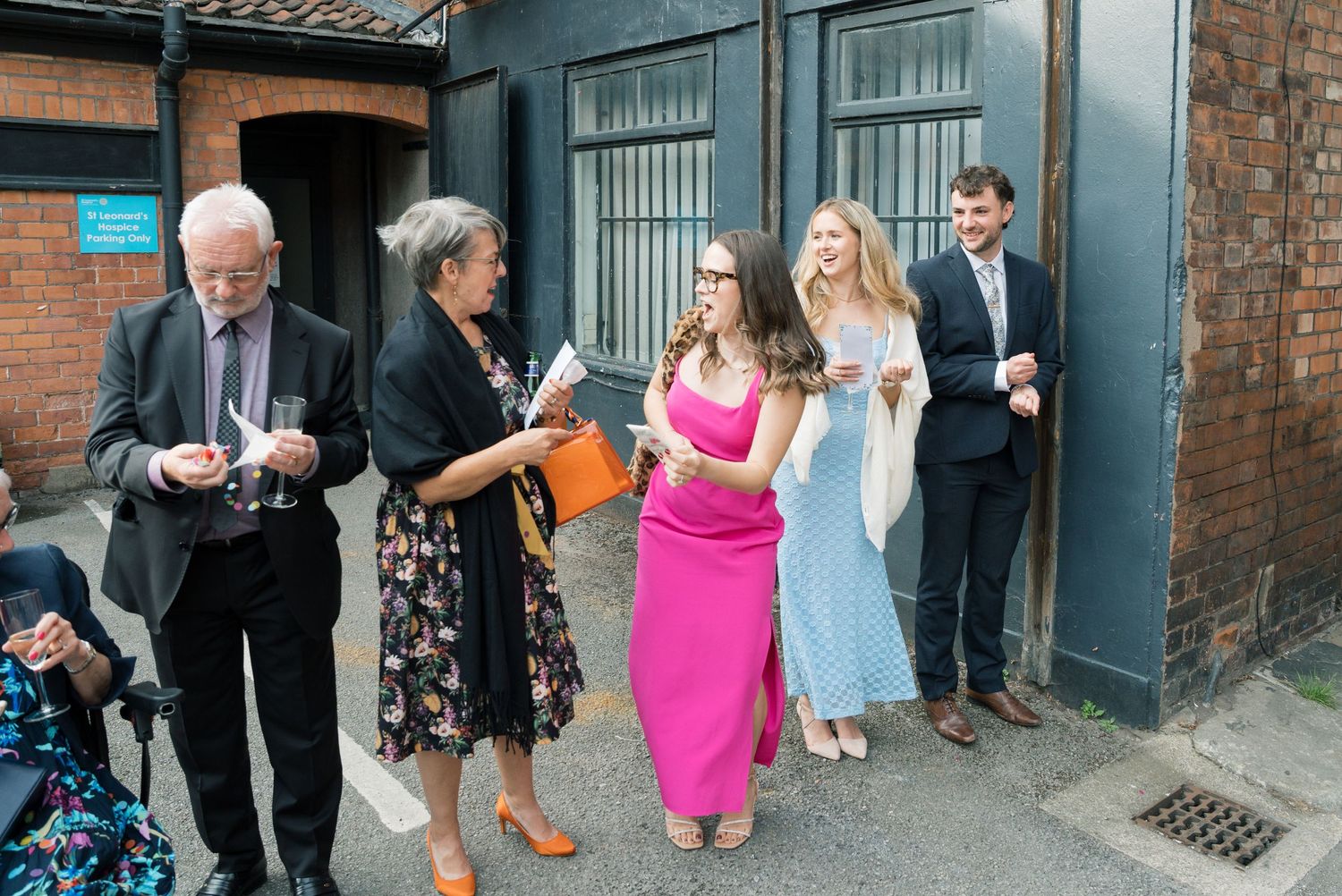Wedding guests in formal attire gathered outside brick building.