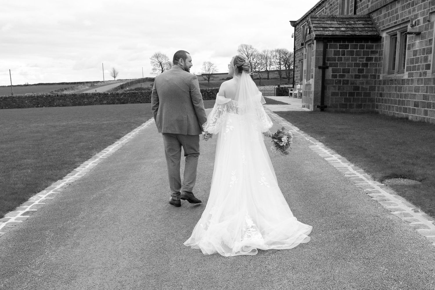 A black and white photo of newlyweds walking down a countryside path near a stone building.