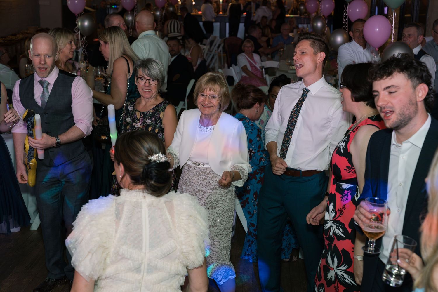 Wedding reception guests dancing and socializing in a dimly lit venue with balloons.