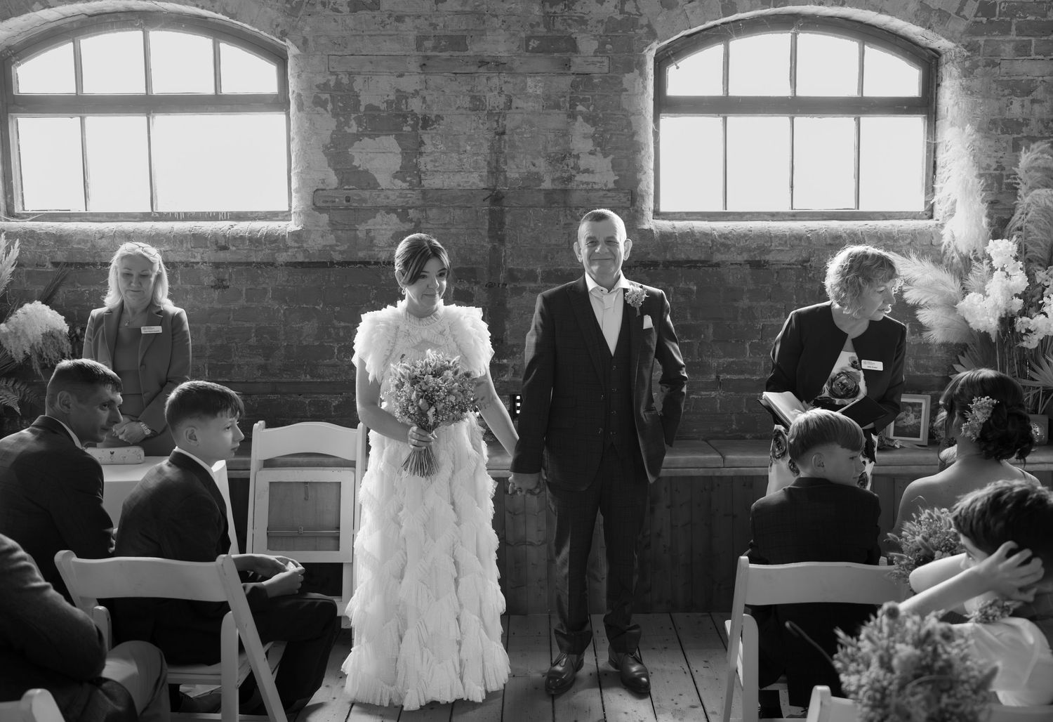 Wedding guests gather in a rustic brick venue with arched windows during a ceremony.