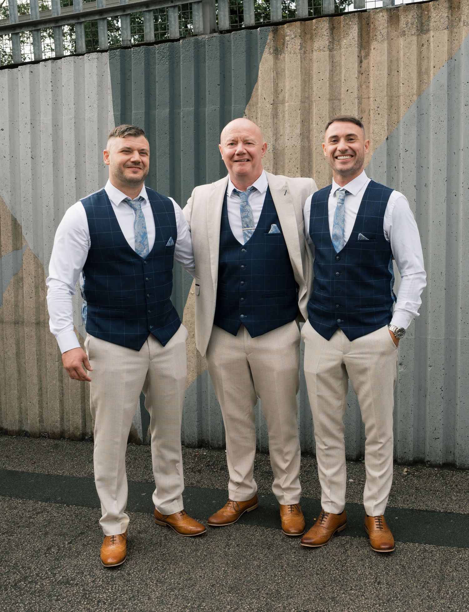 Groomsmen in matching vests and khaki pants stand against industrial wall.