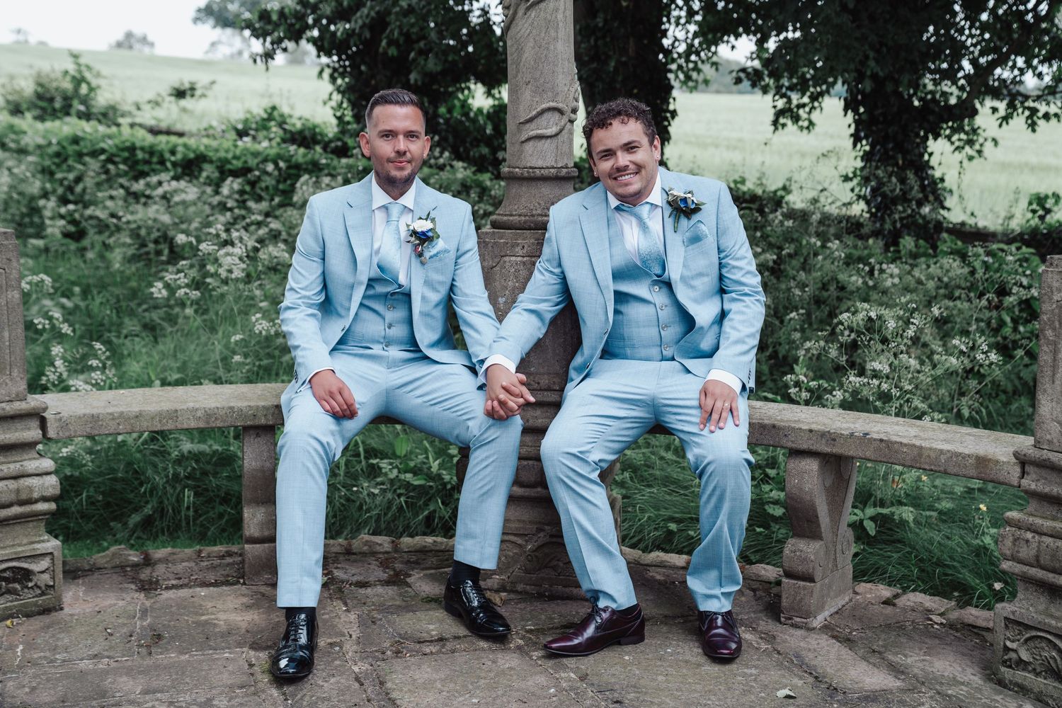 Two grooms in matching light blue suits sit together on stone bench in garden setting.
