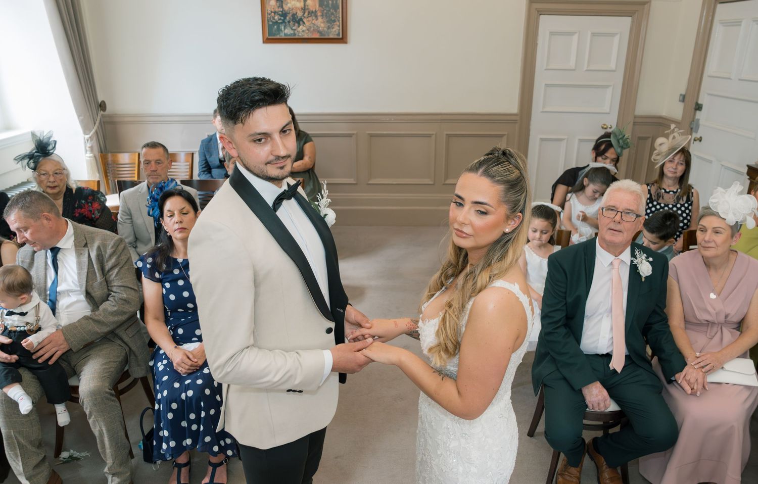 Couple exchanging rings during an indoor wedding ceremony with seated guests in formal attire in the background.