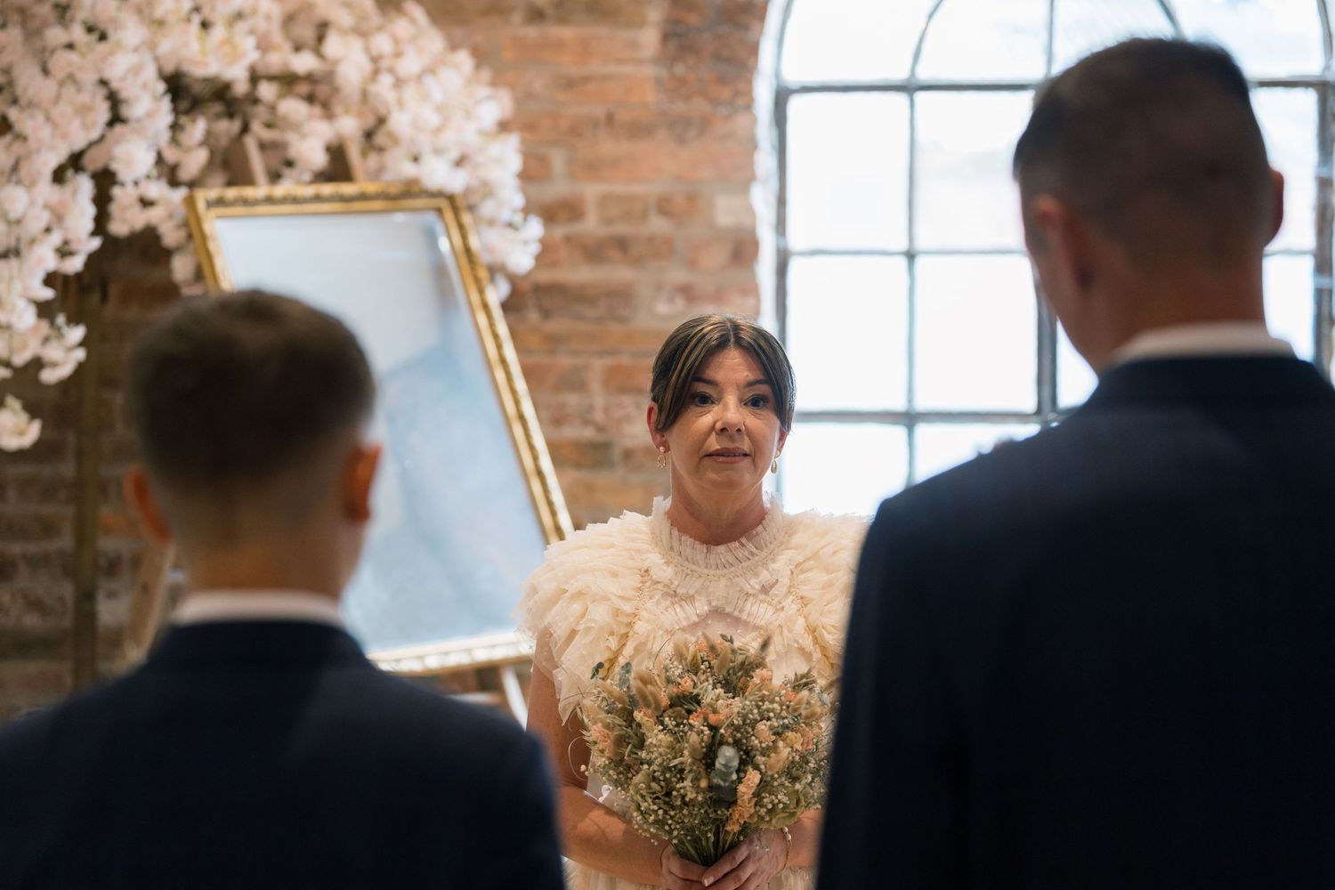 A bride in a lace dress holds a bouquet while standing between two people in dark suits at a rustic indoor venue.