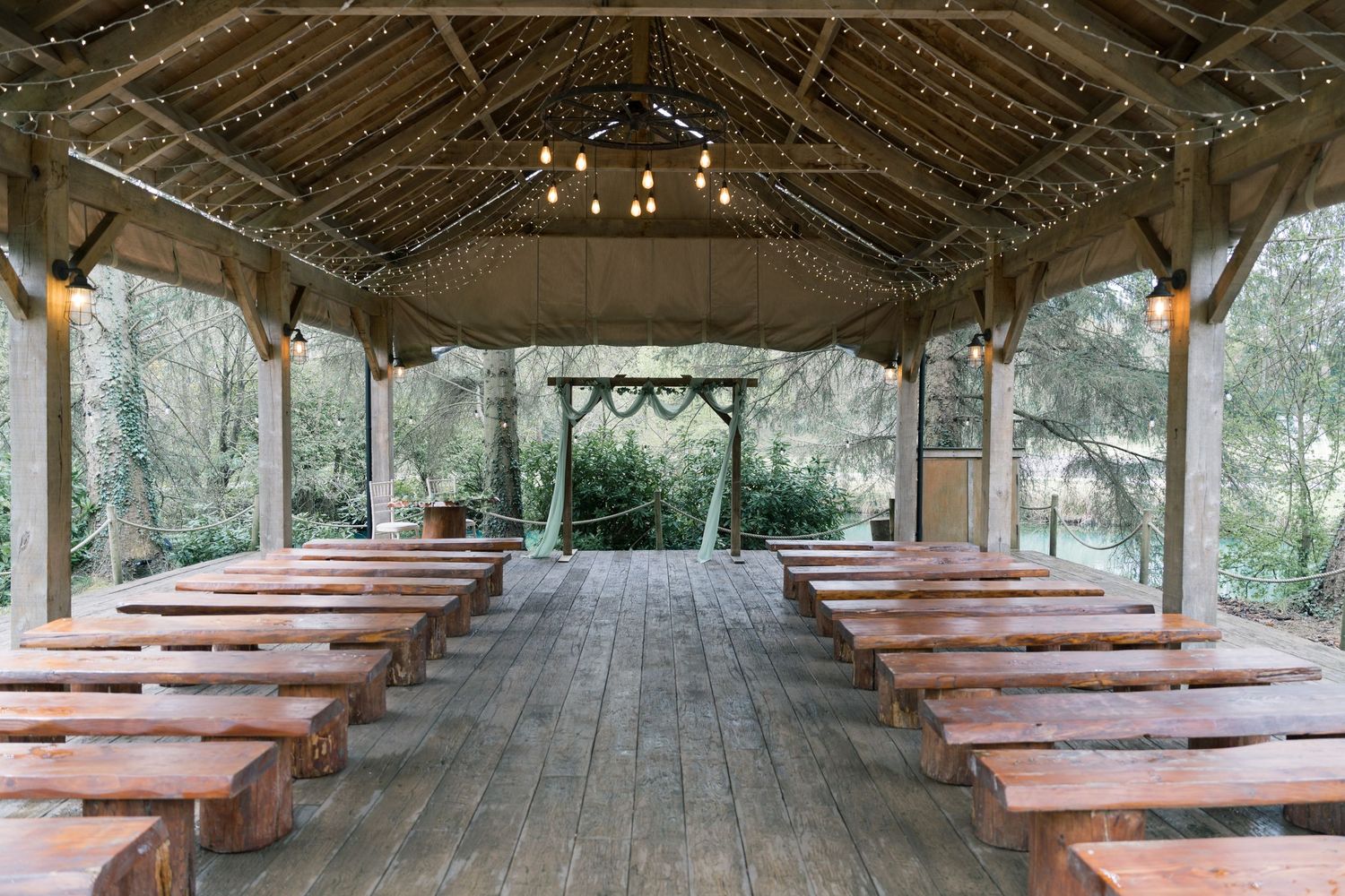 Rustic outdoor wedding venue with wooden benches and string lights under a covered pavilion in a forest setting.