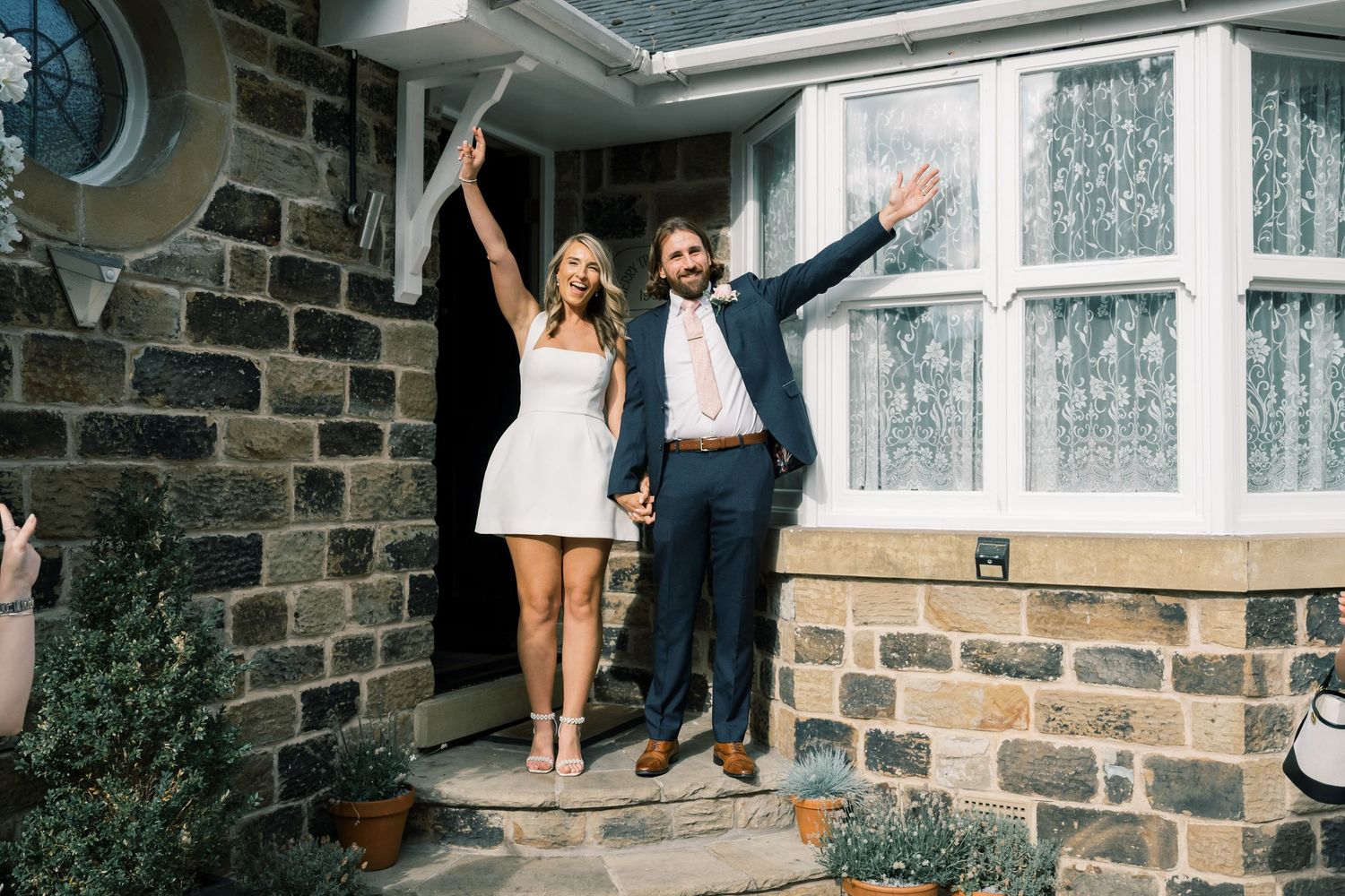 A joyful couple celebrating on stone steps outside a brick house with their arms raised in celebration.