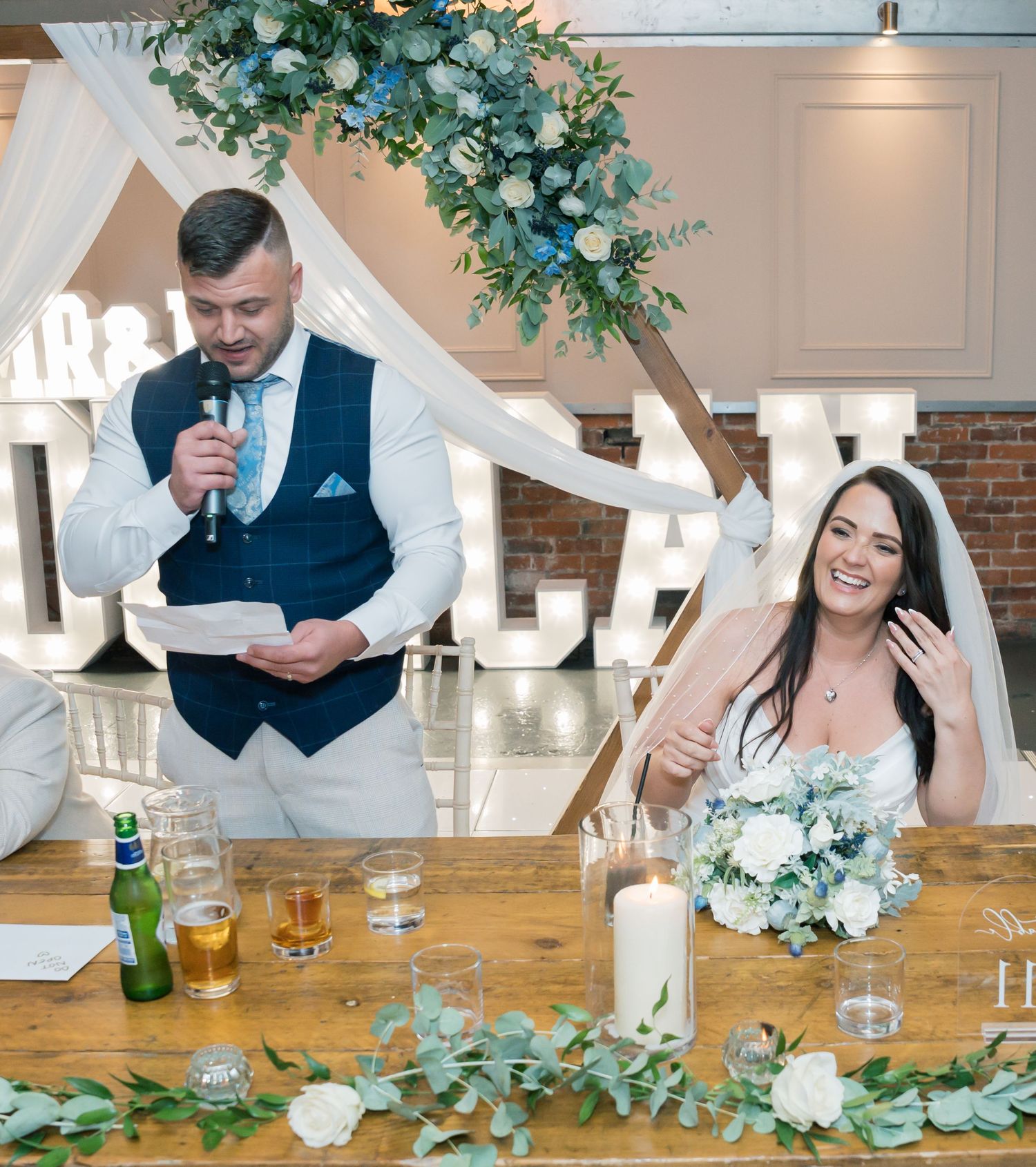 Wedding couple sitting at decorated table with greenery garland and floral centerpieces during reception toast.