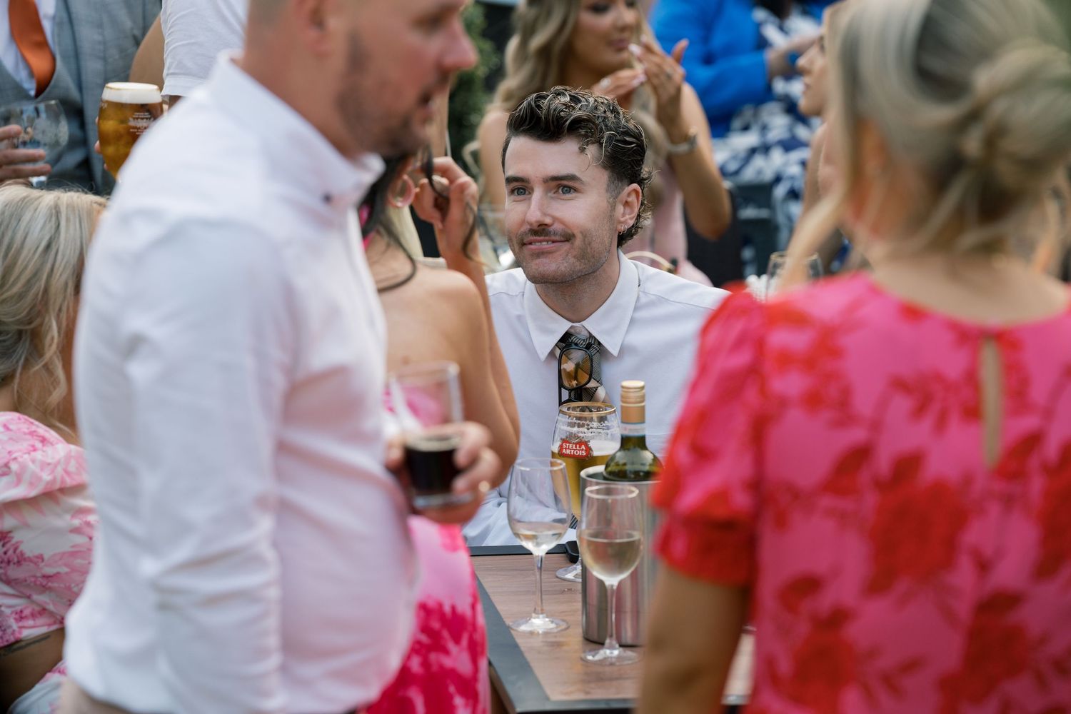 Wedding reception guests mingling at tables with pink and white color scheme.
