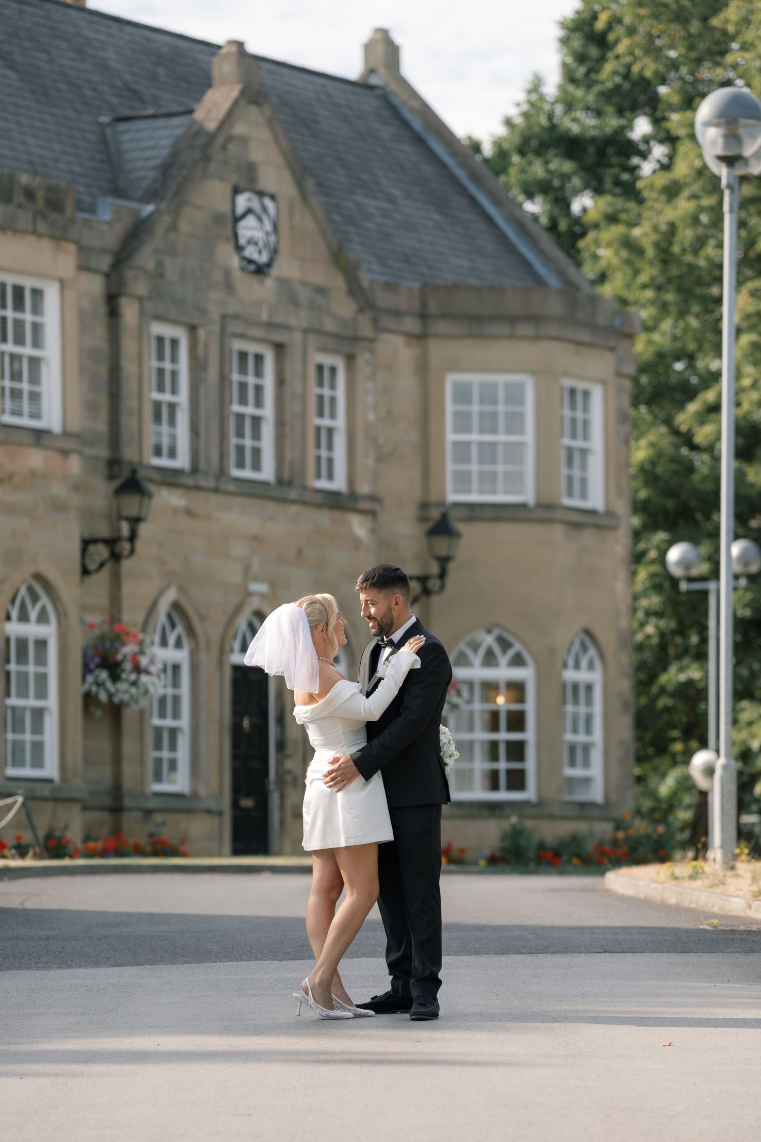 Bride in short white dress and groom in black tuxedo embrace outside a historic stone building on their wedding day.