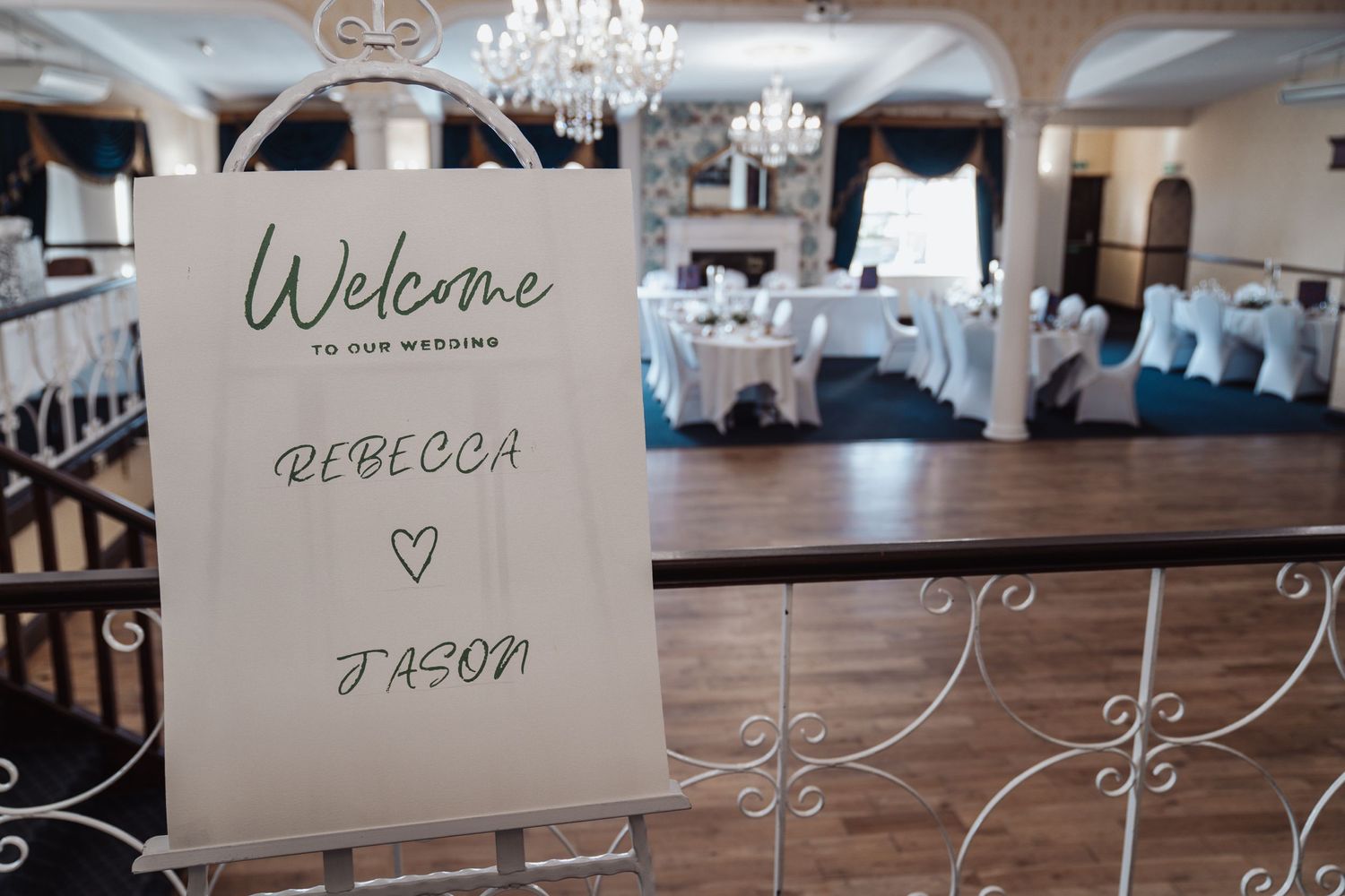 Elegant wedding welcome sign displayed in a ballroom with crystal chandelier and white table settings.