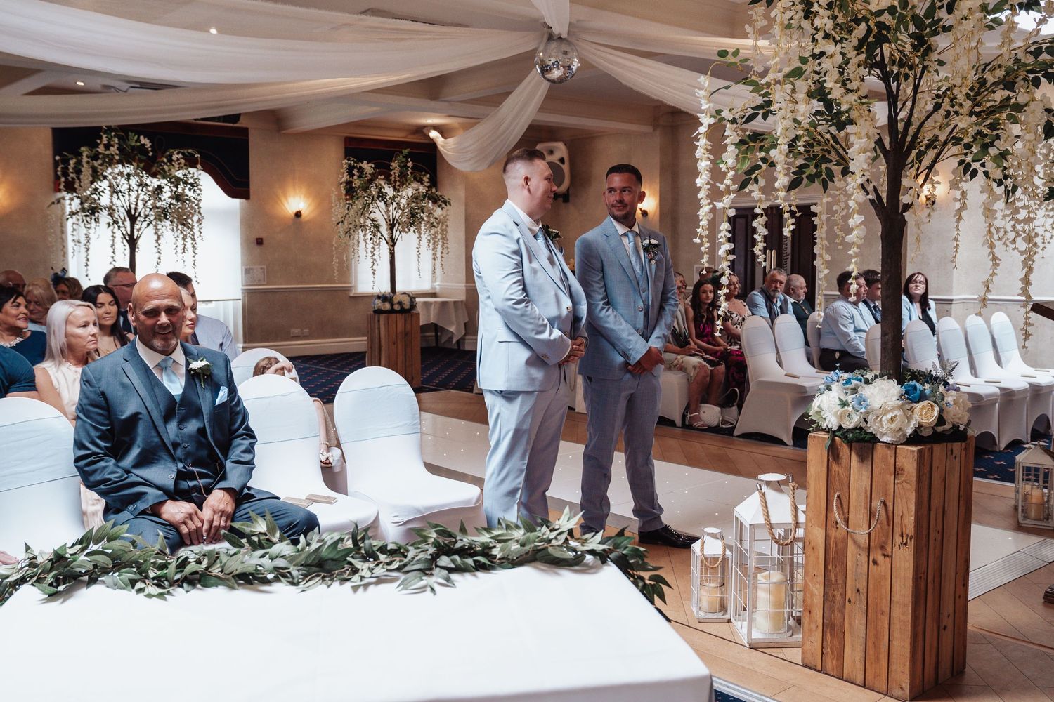 Wedding ceremony setup with blue-suited attendants and white chairs decorated with greenery.