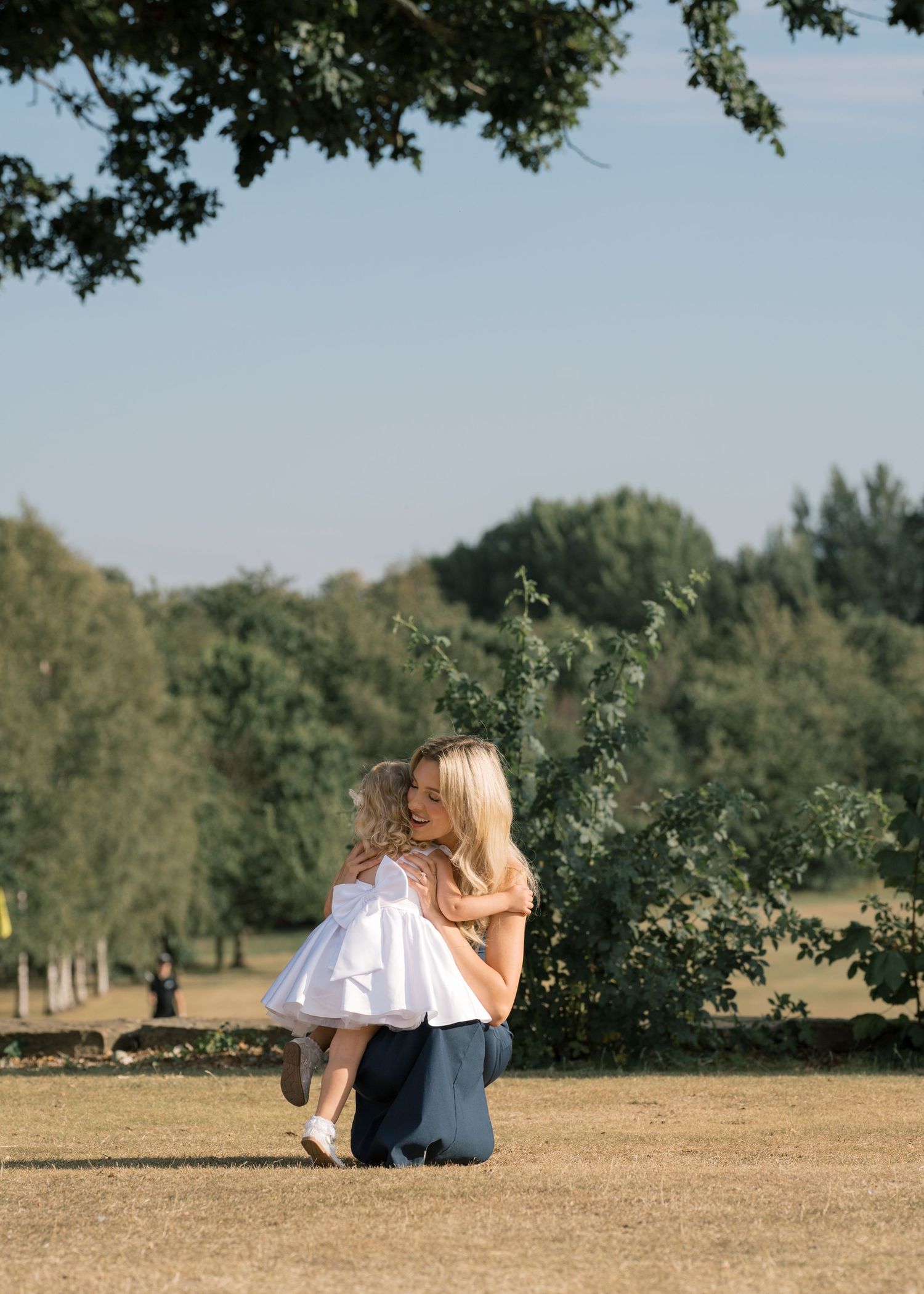 A woman kneels to embrace a young girl in a white dress on a sunny lawn surrounded by lush green trees.