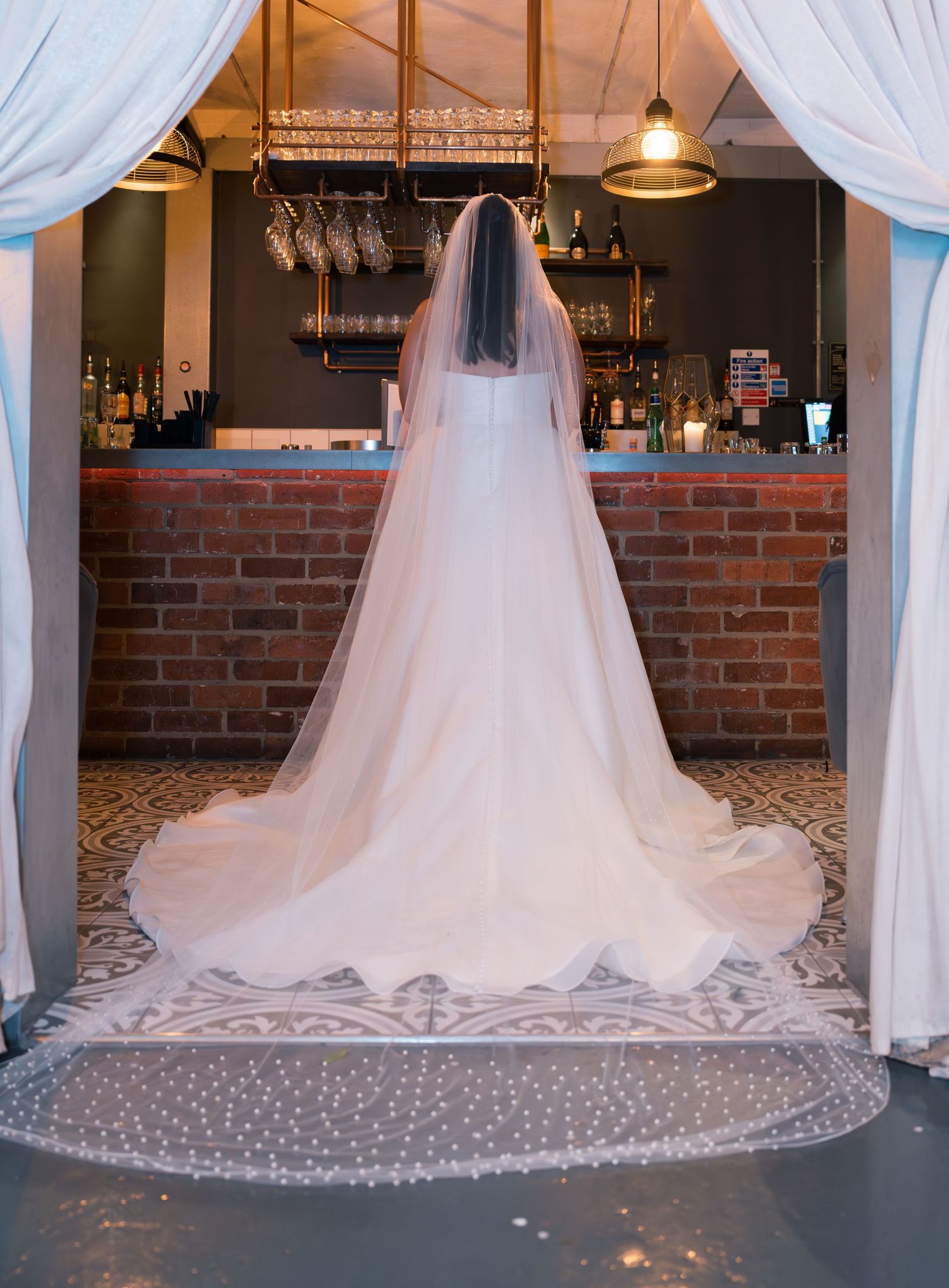 Elegant bridal gown train spread across patterned floor with white drapes and brick wall backdrop.