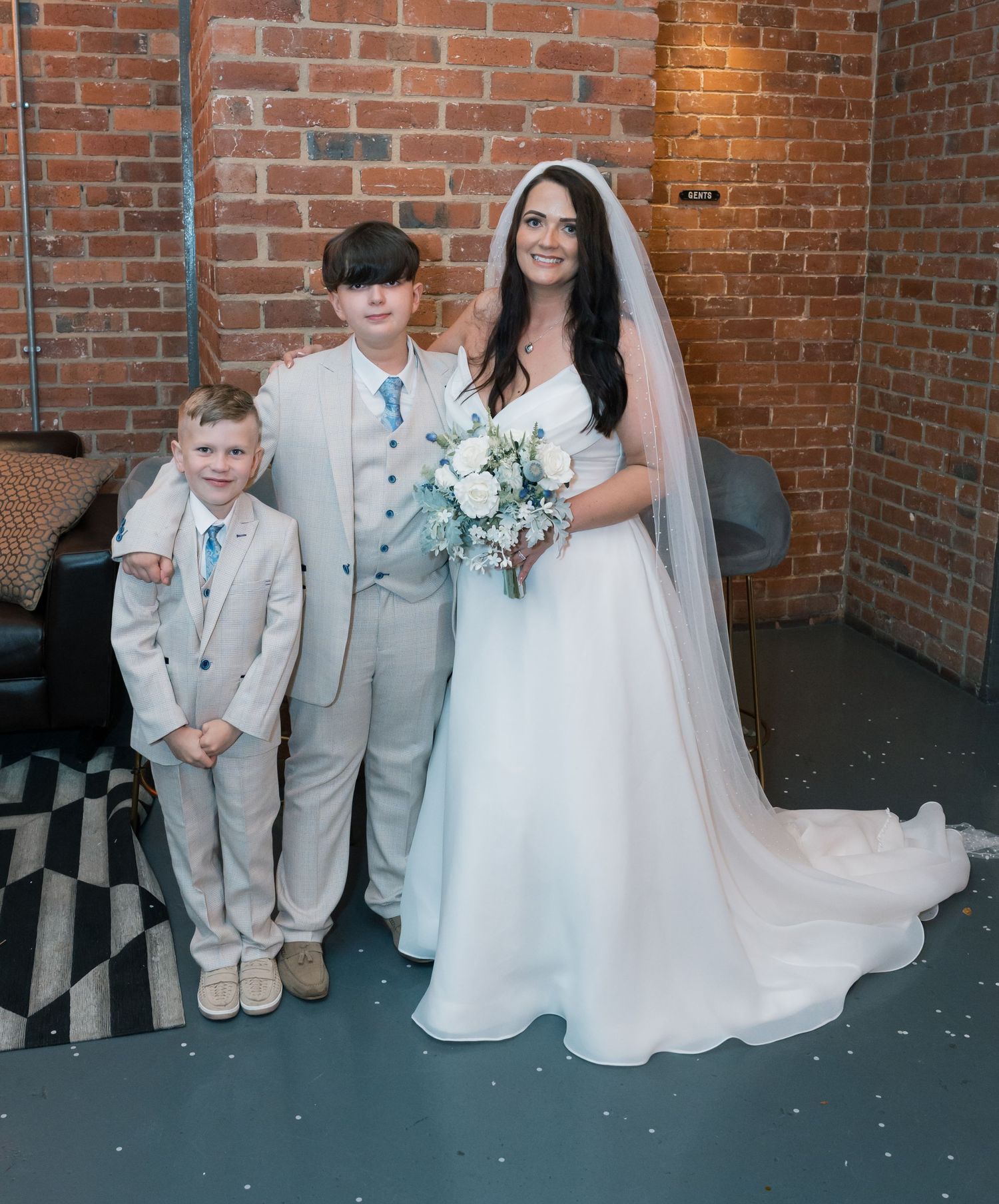 Wedding party poses against brick wall backdrop with bride in white dress and ring bearer in gray suit.