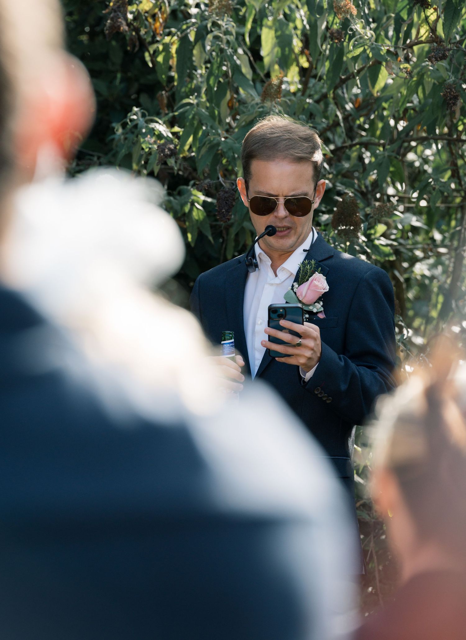 Guest in sunglasses checking phone during outdoor wedding ceremony.