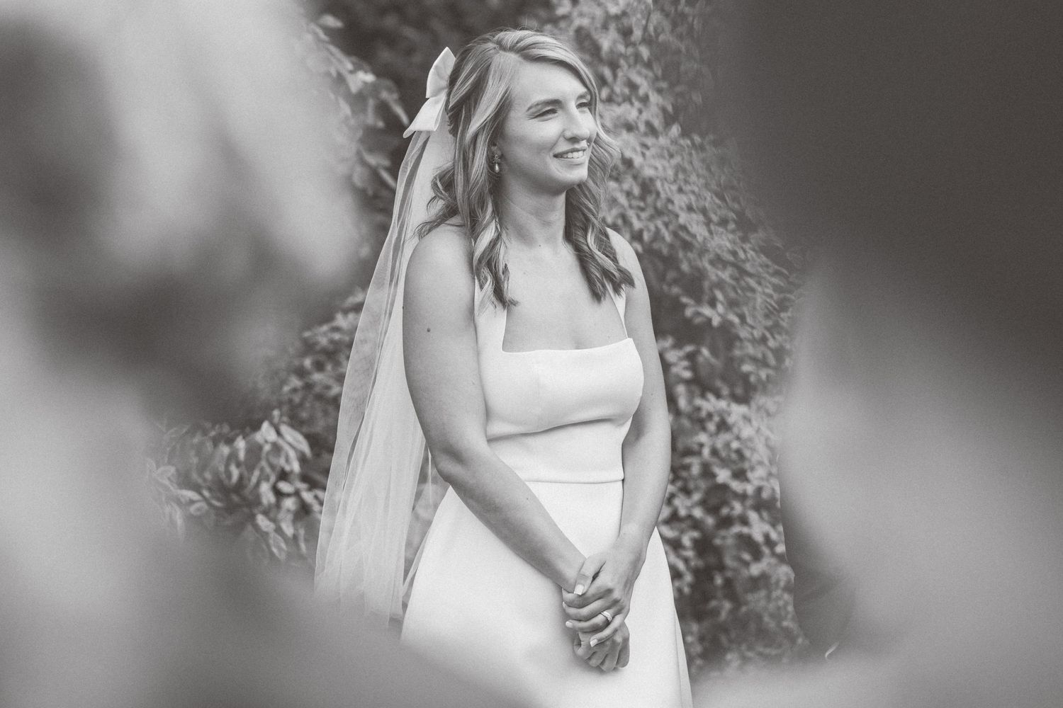Elegant bride in white dress poses against natural outdoor background in black and white photo.