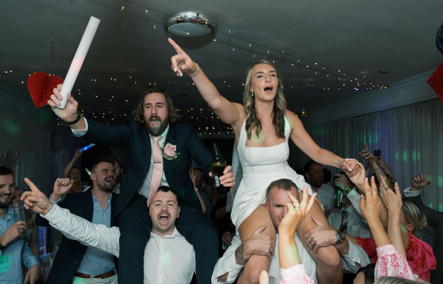 Wedding party guests dance enthusiastically with glow sticks during a nighttime reception celebration.