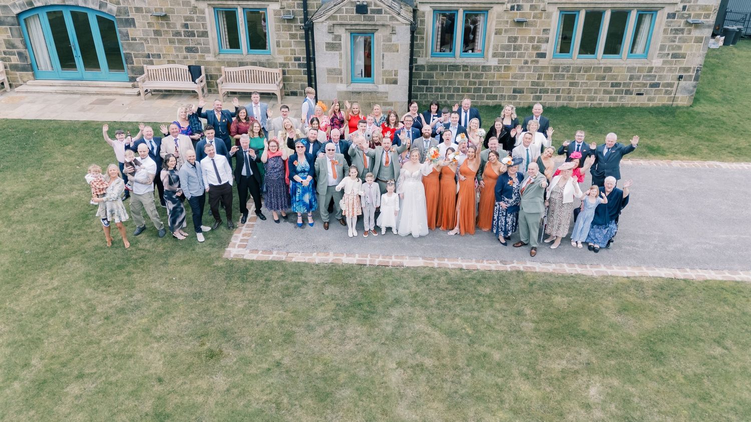 Aerial view of wedding guests gathered outside a stone building for a group photo.