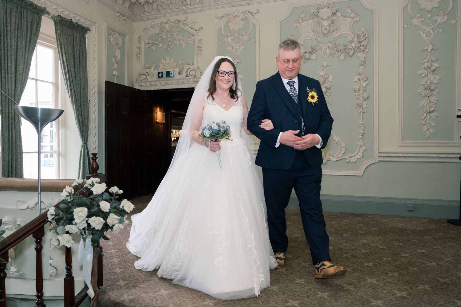 A couple in wedding attire walks through an elegant room with ornate mint green walls and decorative molding.