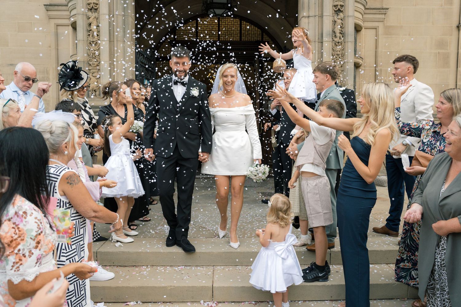 Bride and groom exit church in confetti shower surrounded by smiling wedding guests on sunny day.