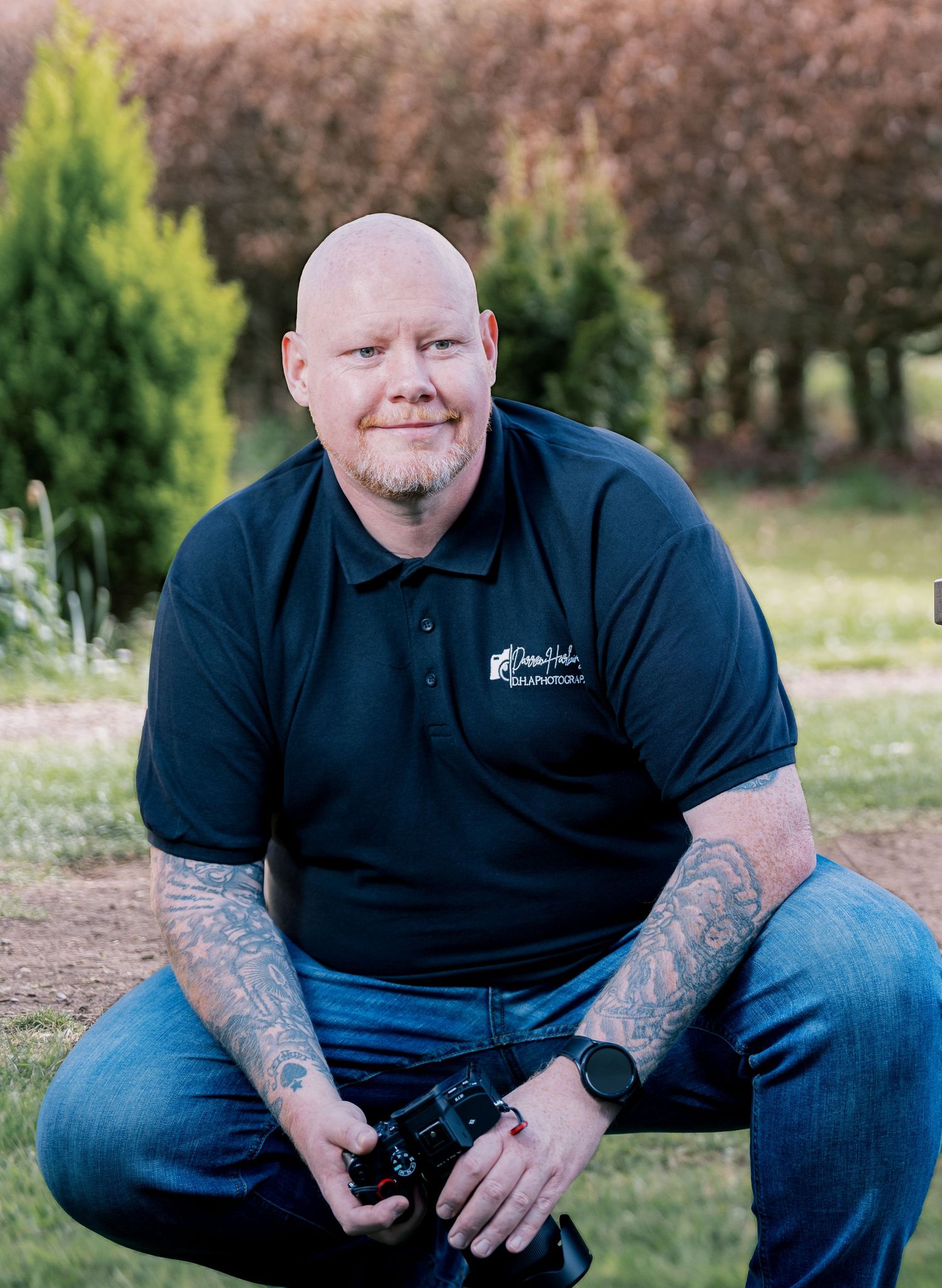 Tattooed bald man crouching outdoors holding a camera, wearing a dark polo shirt and jeans, with trees in background.