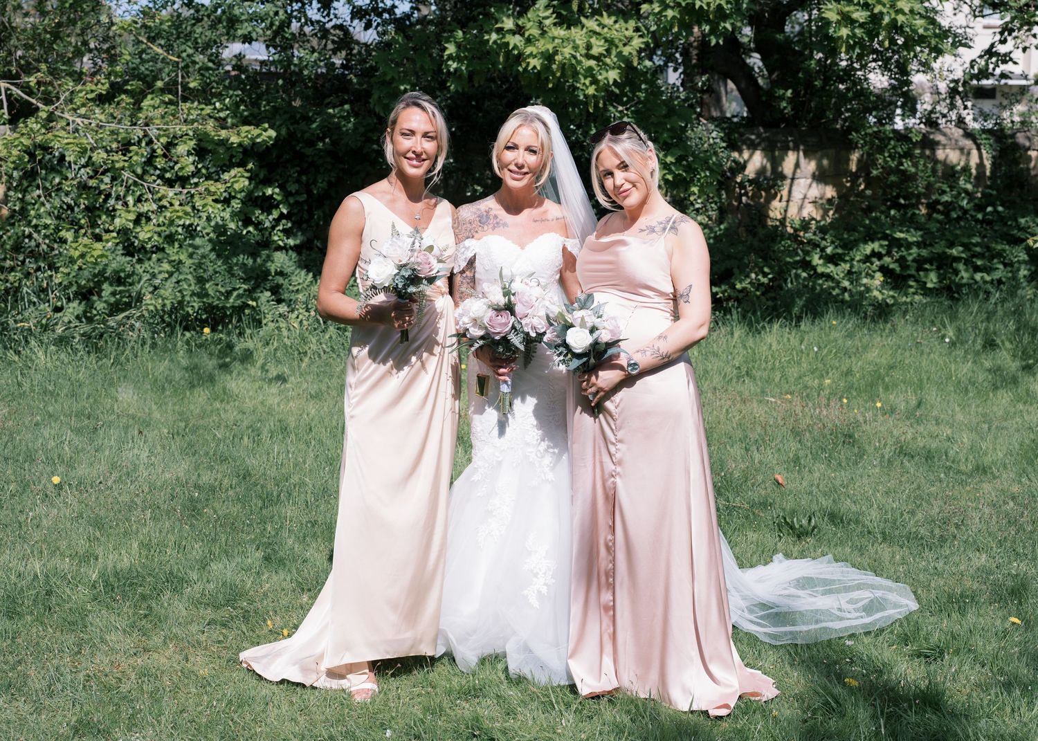 Bride in white gown poses with two bridesmaids in blush pink dresses holding bouquets outdoors.