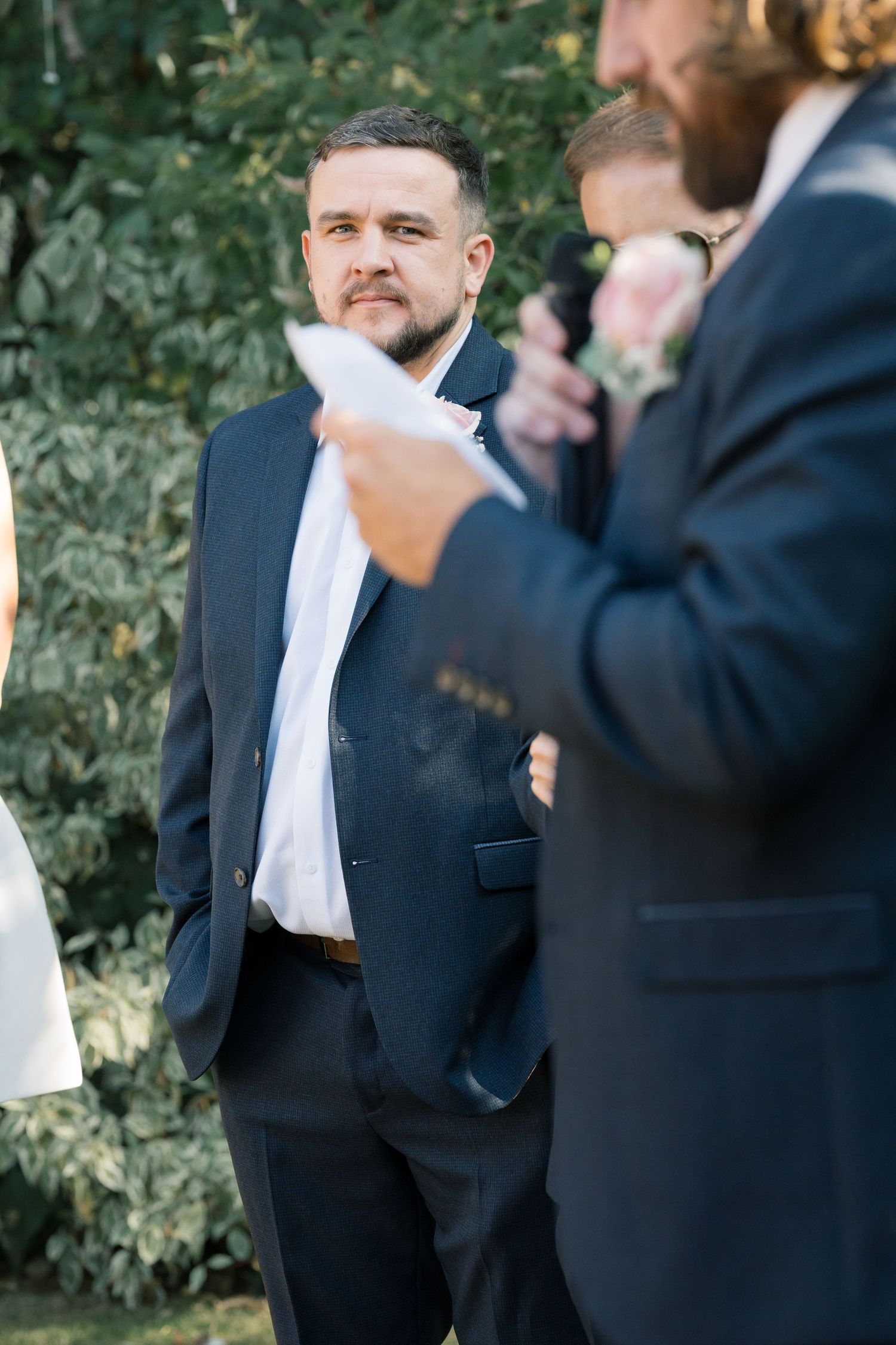 Groomsmen in dark suits stand together during an outdoor wedding.
