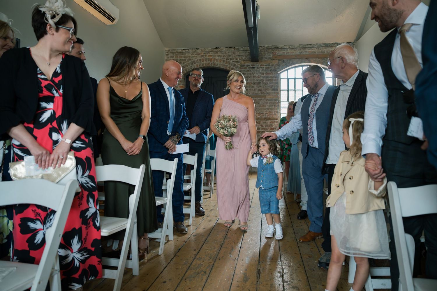 Wedding ceremony guests seated in white chairs inside a rustic brick venue.