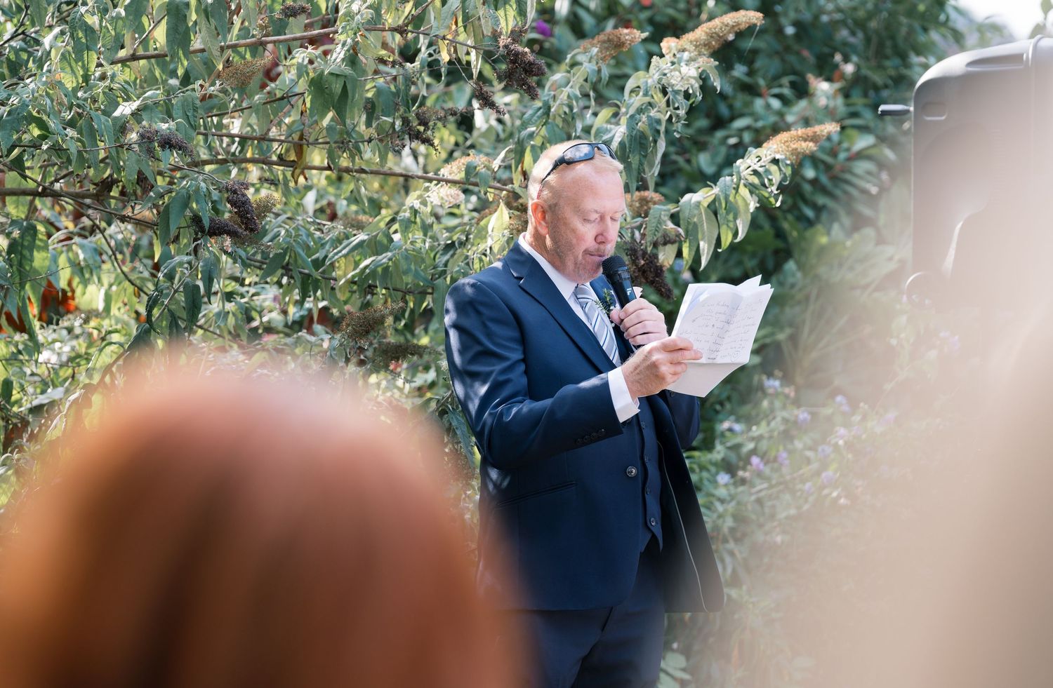 Formal event attendees mingle beneath leafy green trees on a sunny day.