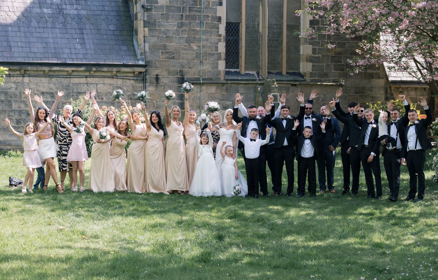 Wedding party celebrating outdoors near a stone church, bridesmaids in champagne gowns and groomsmen in black tuxedos cheering.