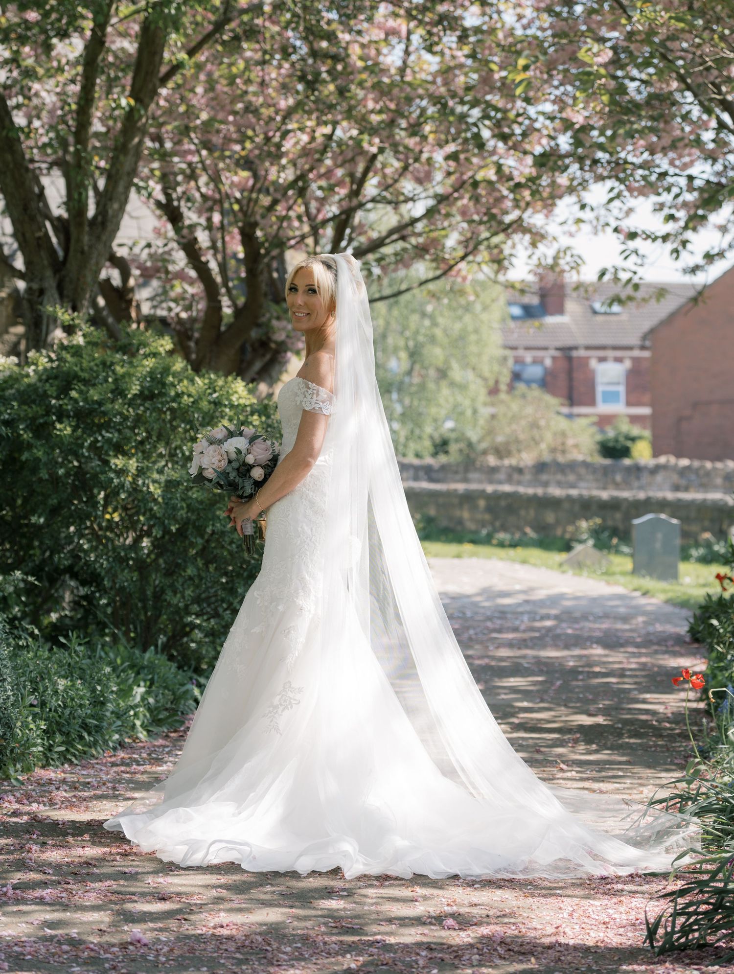 Bride in white off-shoulder gown with cathedral veil holding bouquet, standing on garden path beneath blooming cherry blossom tree.