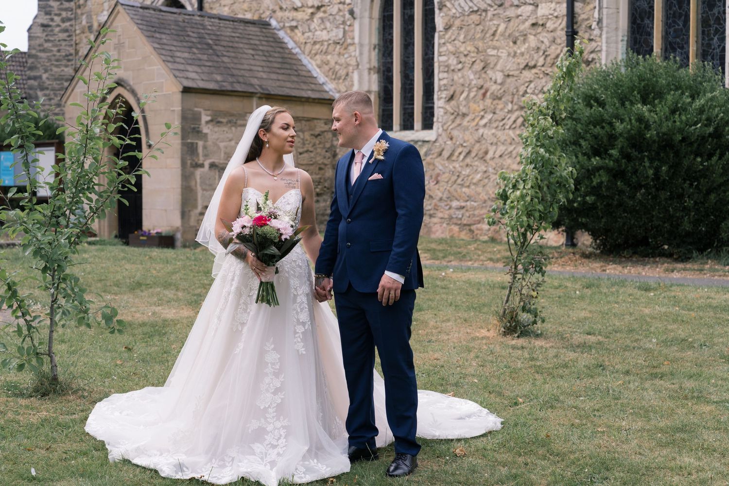 An elegant couple stands on a church lawn, with the bride in white lace and groom in navy blue suit holding a bouquet.