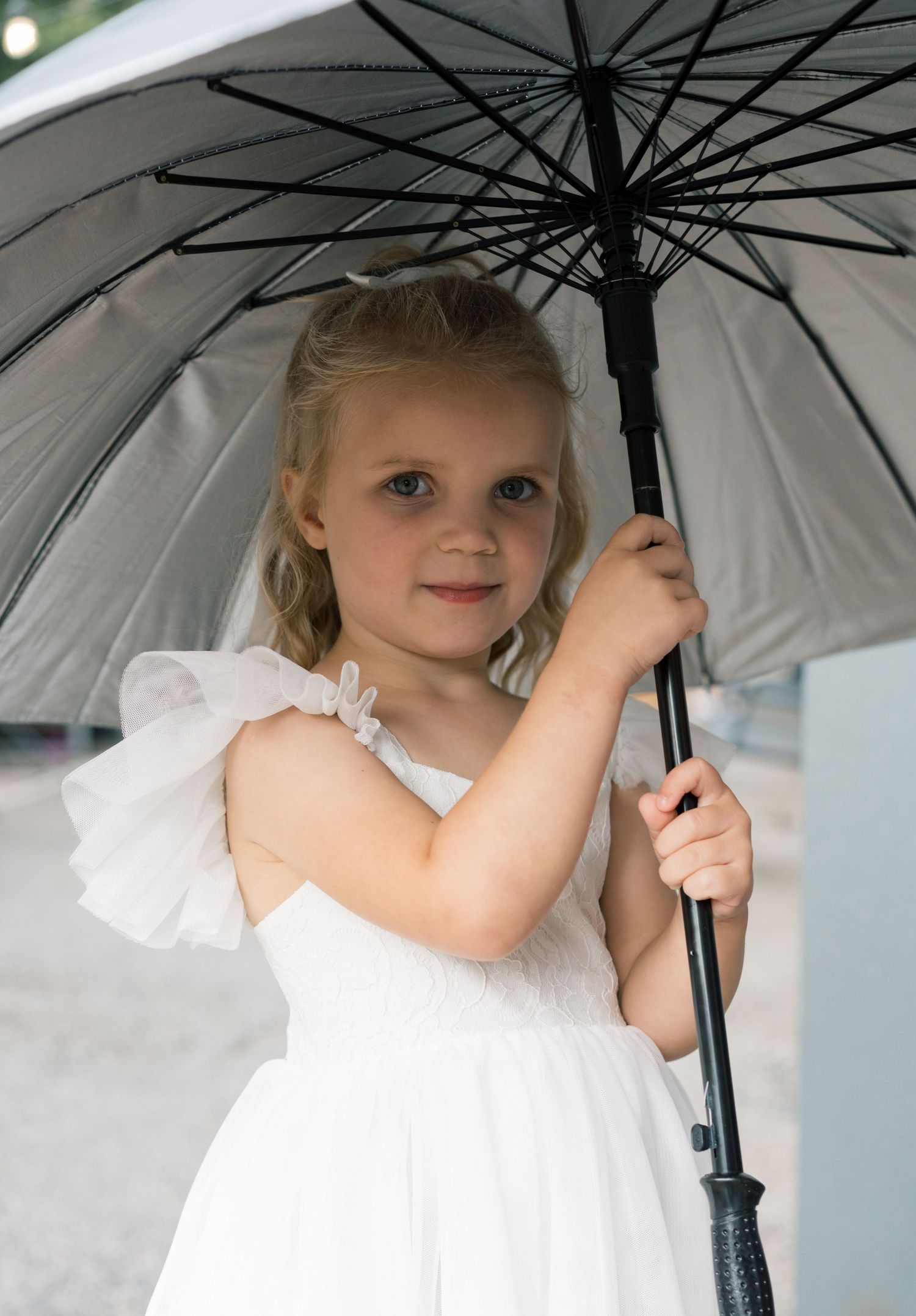 Young attendant in white dress holds white umbrella during outdoor wedding ceremony.