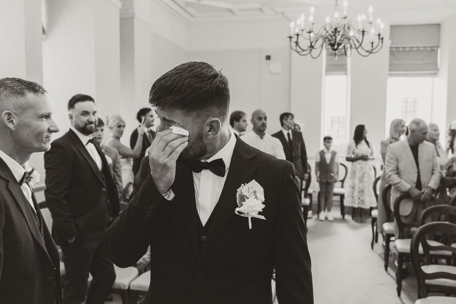 Emotional groom wiping tears at wedding ceremony in elegant venue, wearing black tuxedo with white boutonniere.