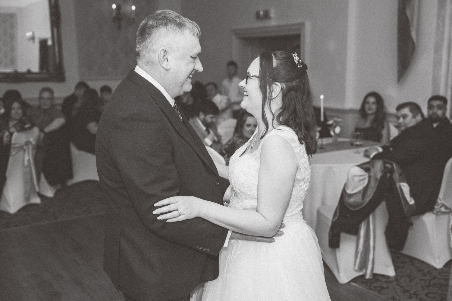 A black and white photograph of a couple sharing a dance together at a formal event with guests seated in the background.