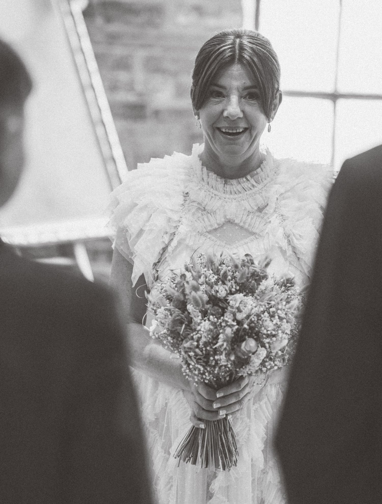A bride in a white ruffled dress holds a delicate bouquet of flowers while smiling during her wedding day.