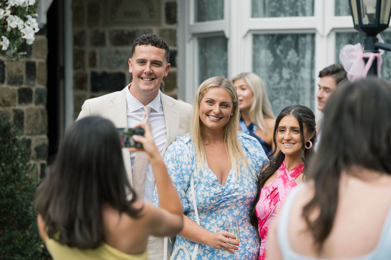 Group of wedding guests mingling outdoors in front of stone building.