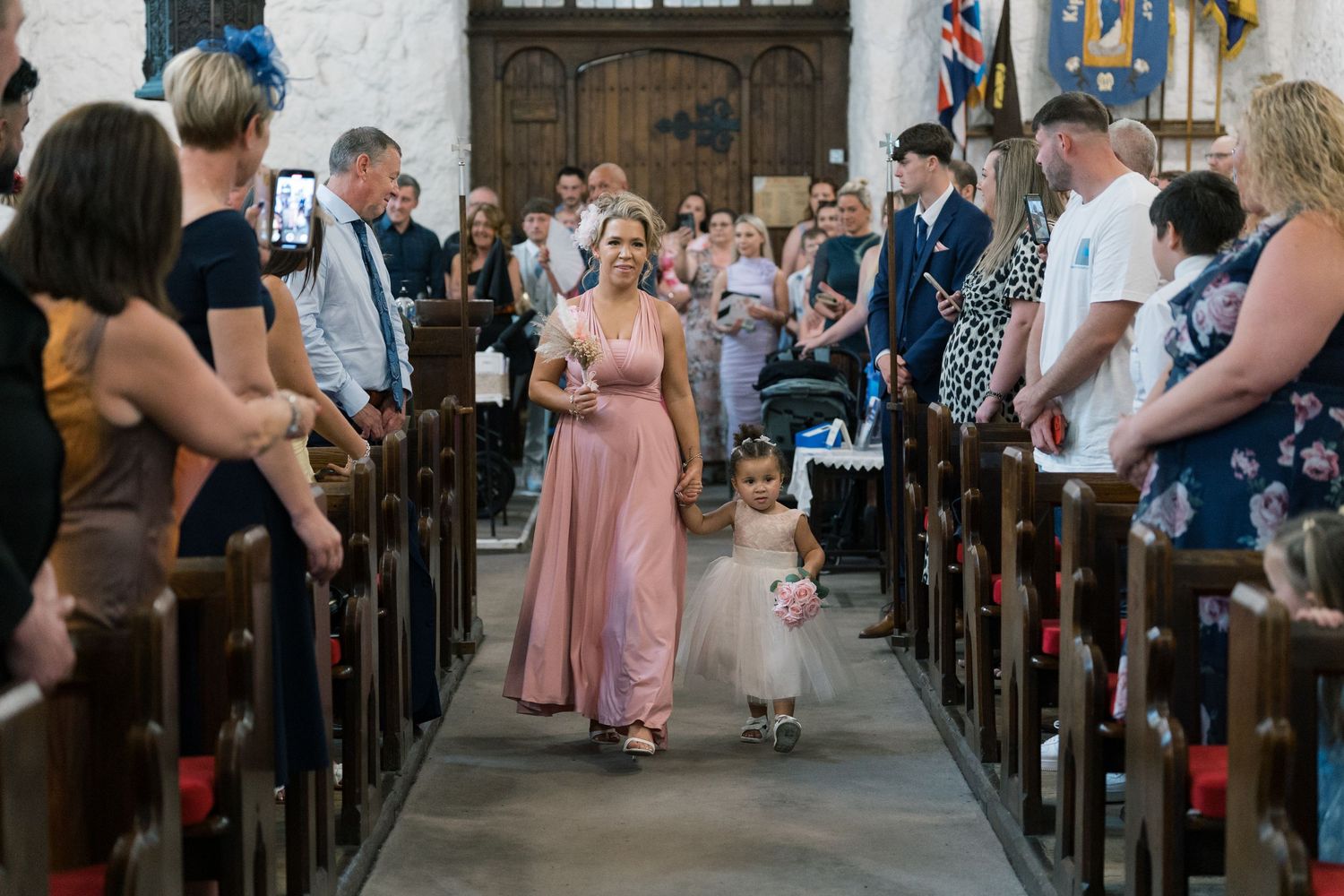 Wedding guests watch as bridesmaids walk down the church aisle with a small flower girl in a white dress.