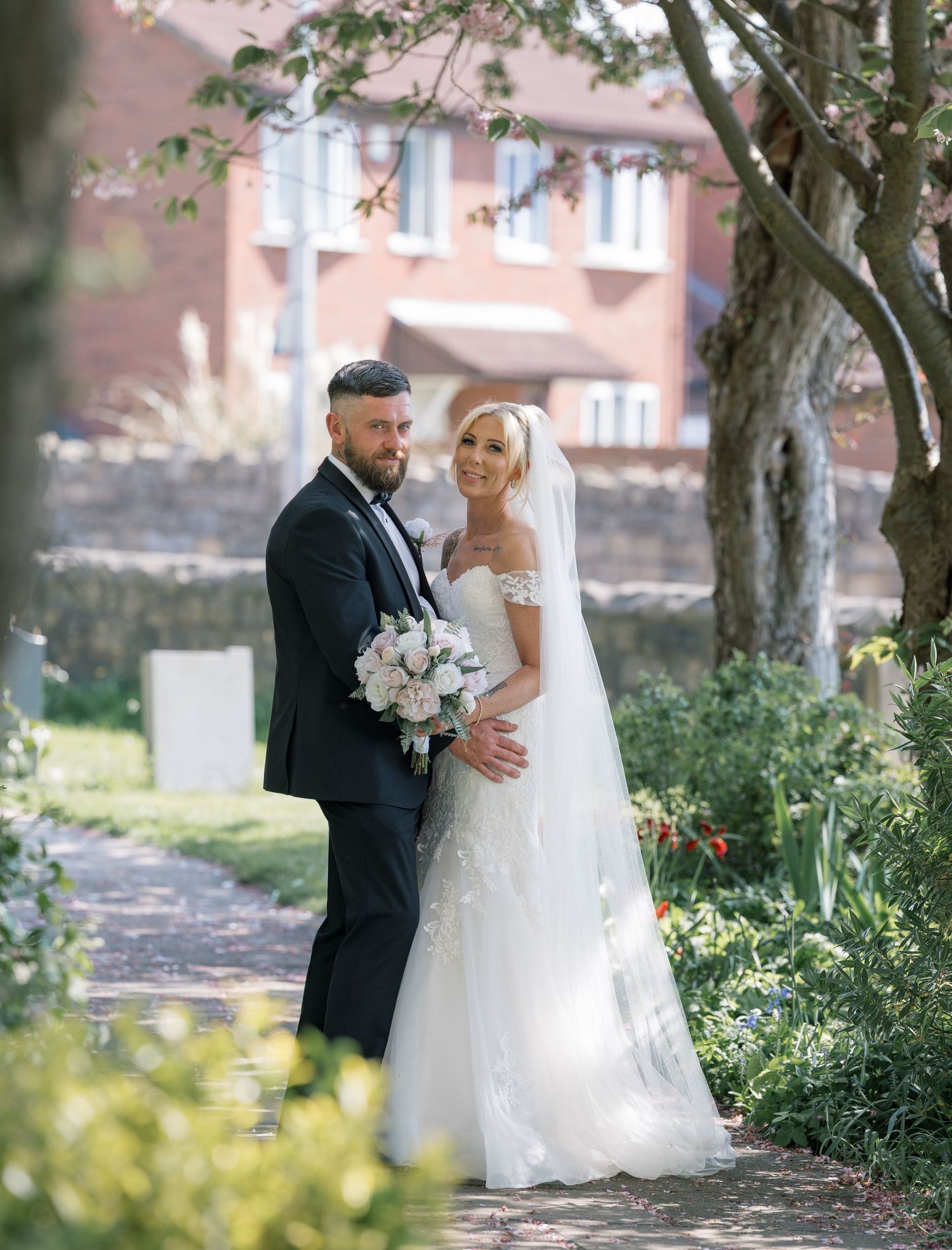 A bride in an off-shoulder lace gown holding white bouquet stands with a groom in a dark tuxedo in a church grounds in Leeds.