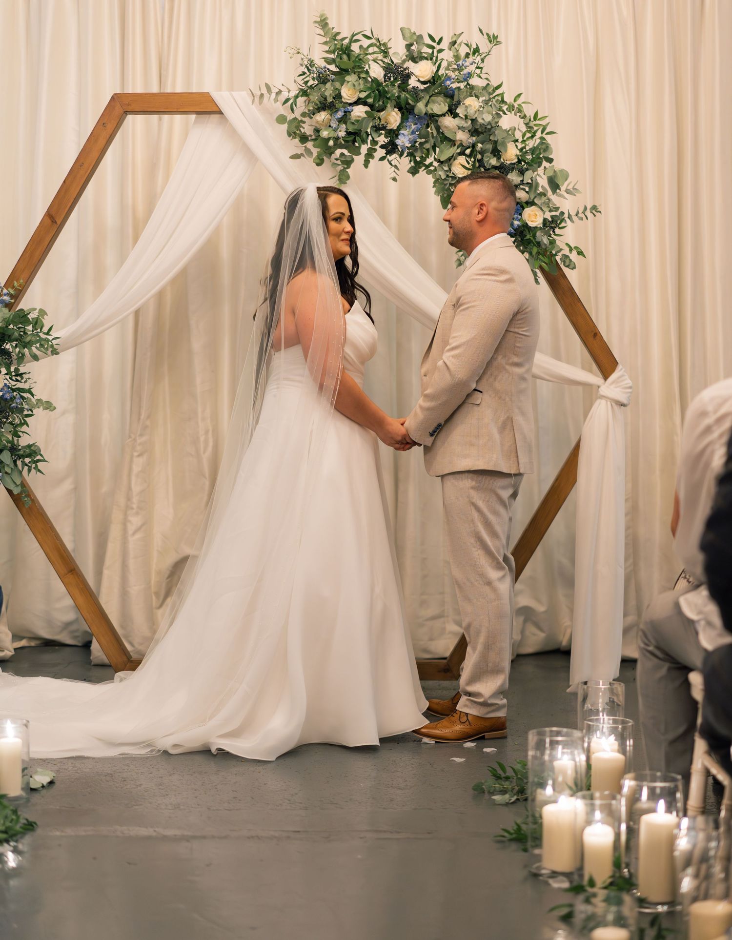 Wedding couple shares an intimate moment under a wooden arch decorated with greenery and white drapes.