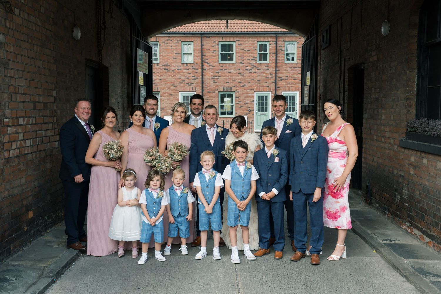 Wedding party poses for group photos under brick archway at historic venue.