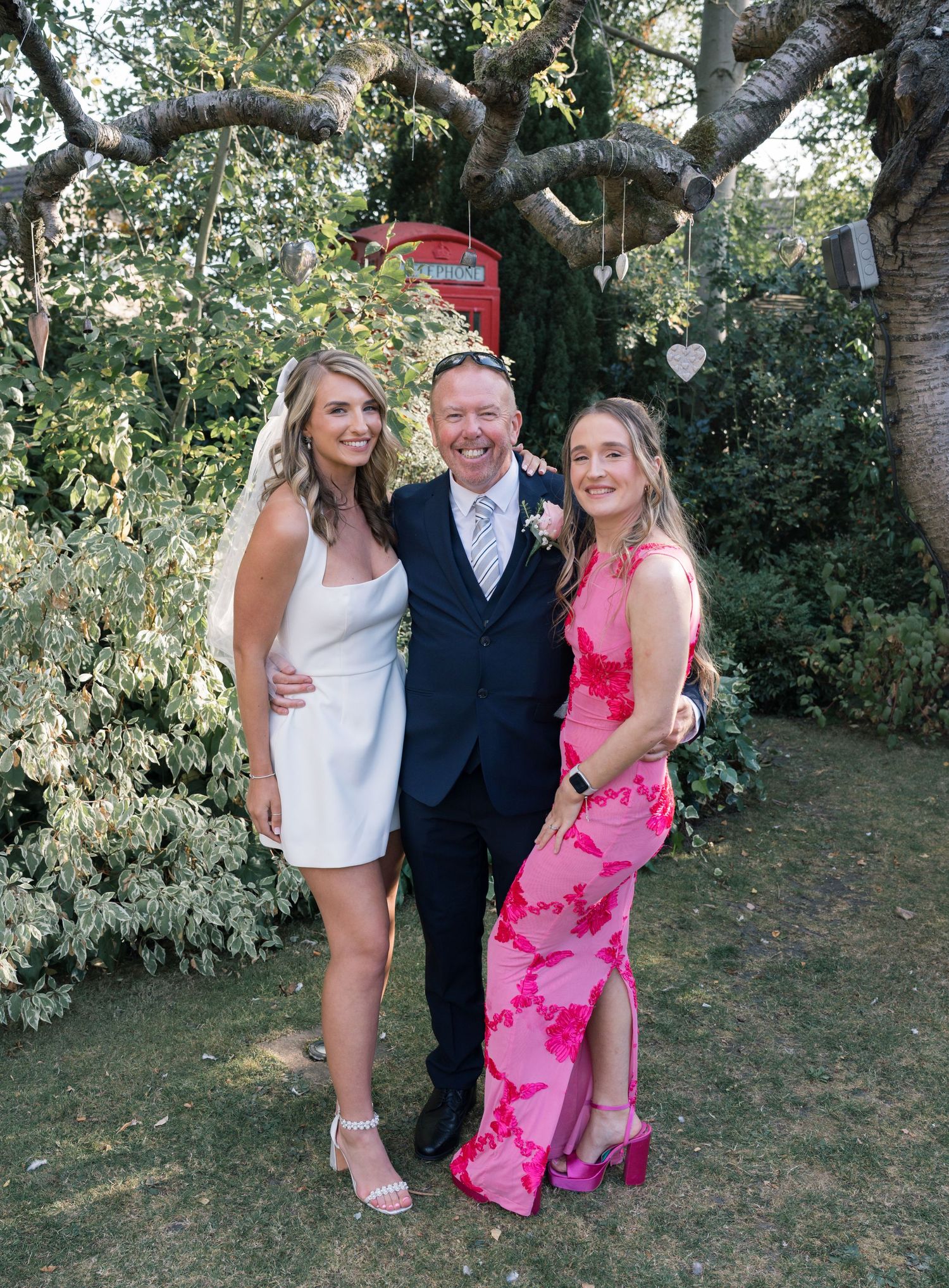 Group photo in a garden setting with people wearing white, black and pink outfits under large trees.