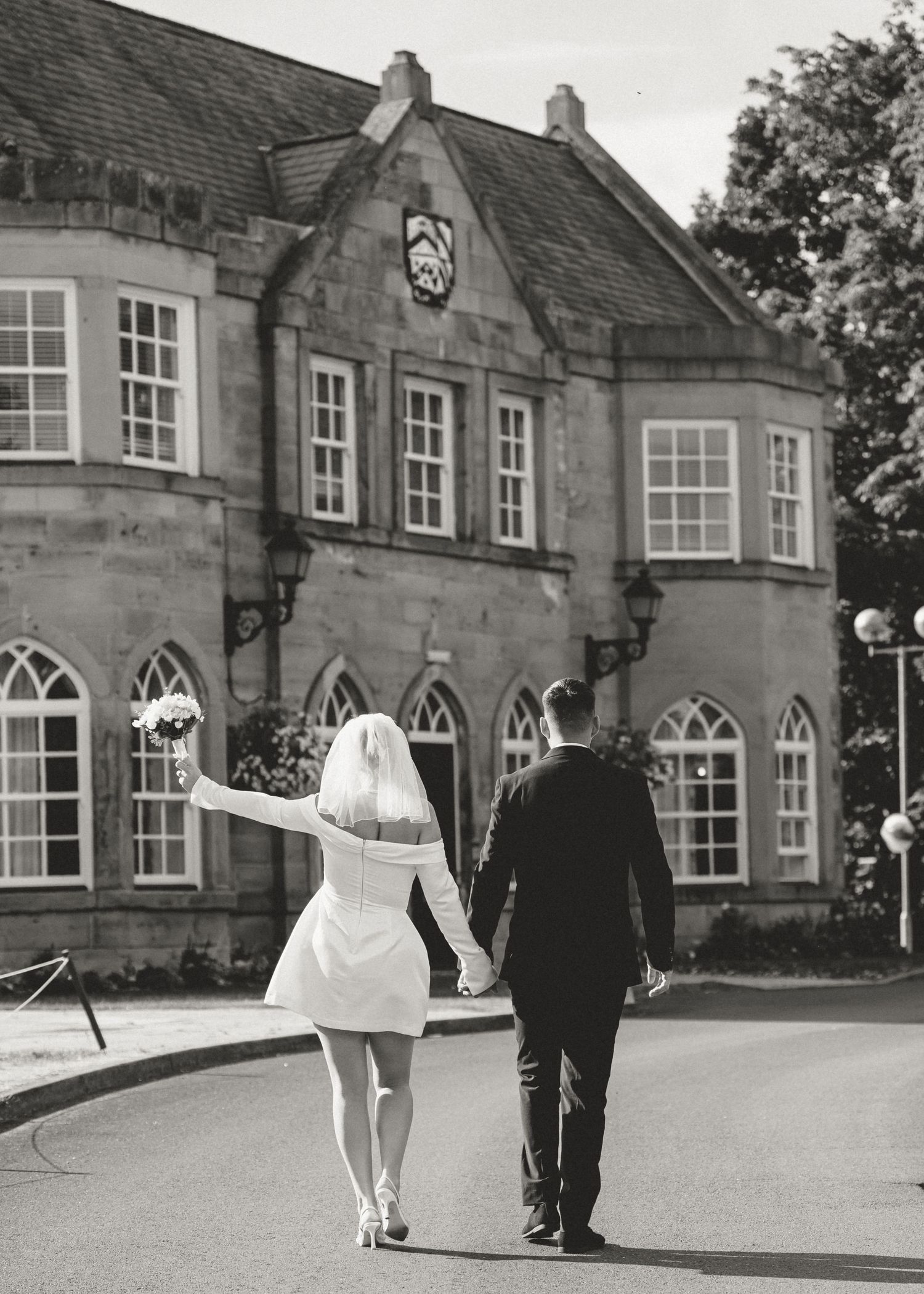 Black and white photo of a bride and groom walking hand in hand toward an elegant stone building.